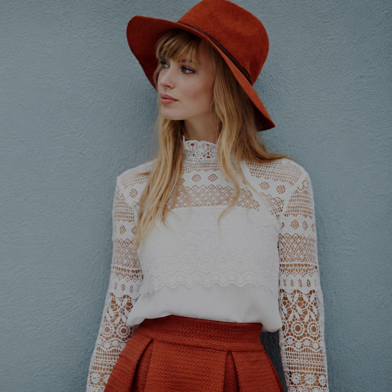 A woman wearing a wide-brimmed rust-orange hat and a white lace long-sleeve top, standing against a textured light blue-gray wall.