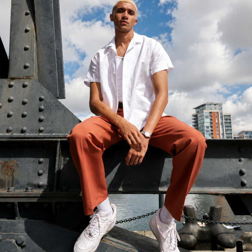 A man posing outdoors wearing a white button-up shirt and rust pants