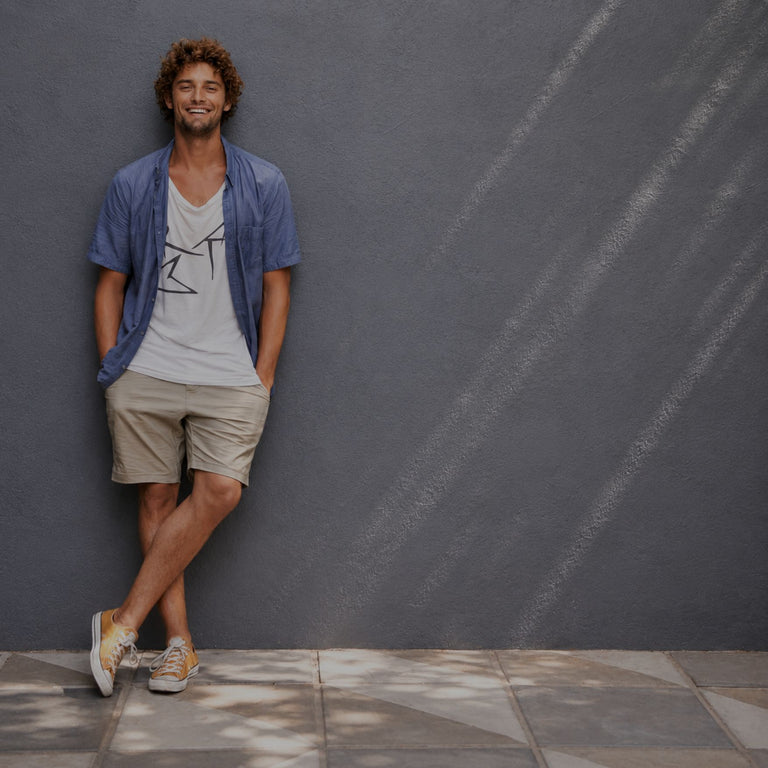 A smiling man with curly brown hair stands against a textured gray wall, wearing a light blue open button-up shirt over a white graphic t-shirt and beige shorts.