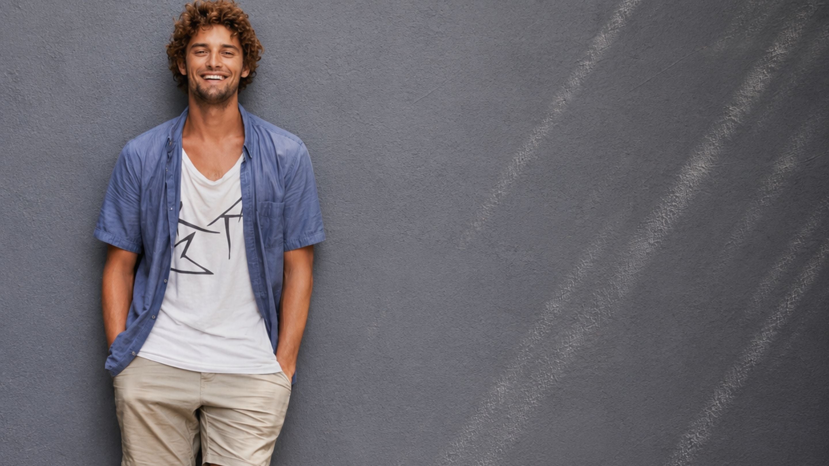 A smiling man with curly brown hair stands against a textured gray wall, wearing a light blue open button-up shirt over a white graphic t-shirt and beige shorts.
