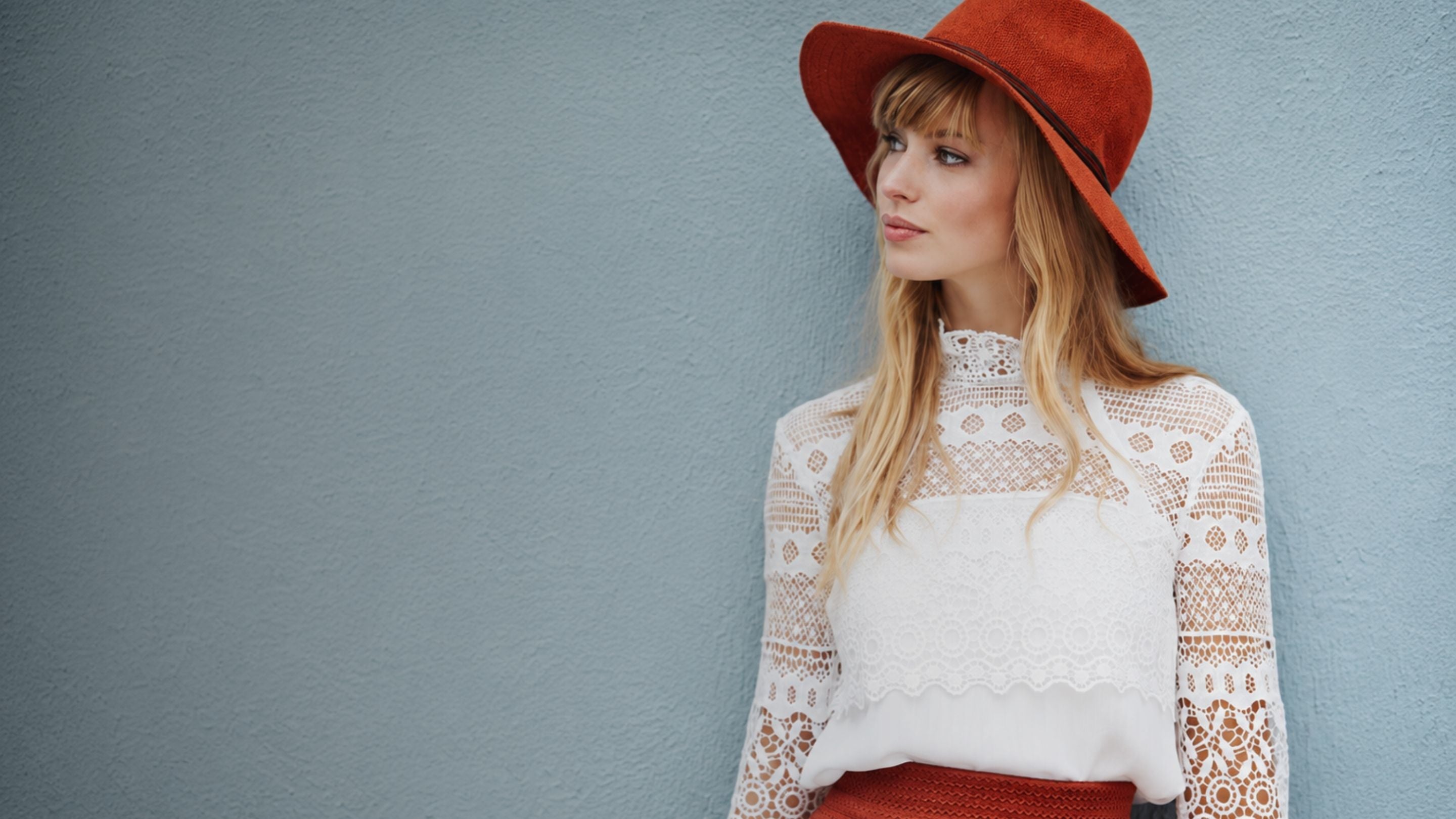 A woman wearing a wide-brimmed rust-orange hat and a white lace long-sleeve top, standing against a textured light blue-gray wall.